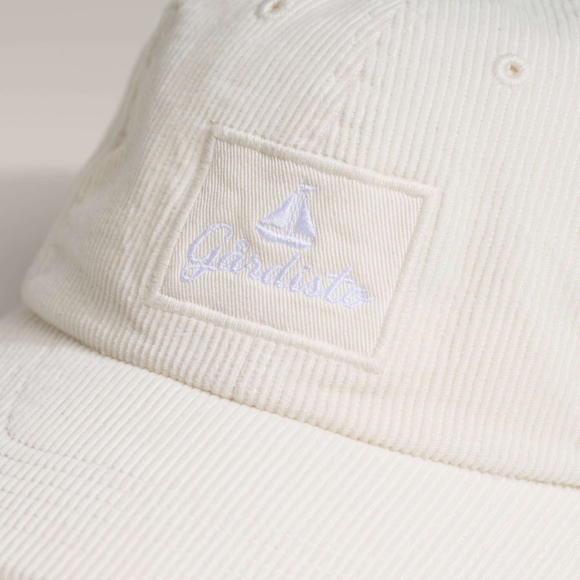 Brodert Gårdisto-logo med seilbåt på sandfarget cordfløyelscaps