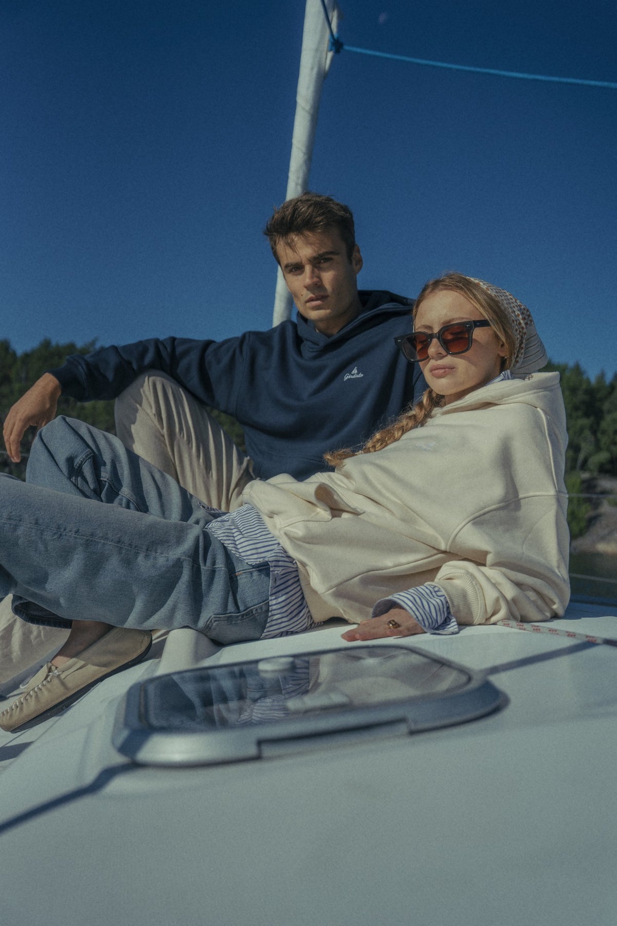 Male model in a navy Gårdisto hoodie and female model in a cream Gårdisto hoodie relaxing on a sailboat.